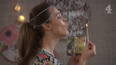 An animated GIF of a women with strings of jewels around her head blowing out the flame of a stick of incense.
